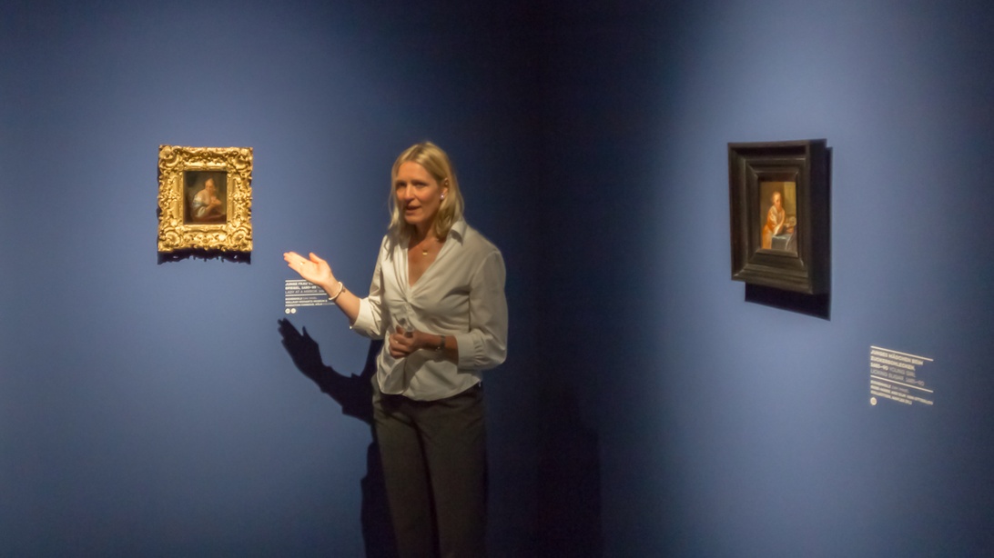 En professionelt klædt kvinde præsenterer i et kunstgalleri og peger mod klassiske malerier på en blå væg, hvilket demonstrerer galleriets forretningsdrift.